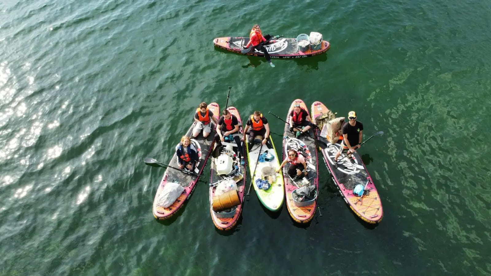 Vu du ciel, des bénévoles en gilet de sauvetage sur leurs stand up paddles chargés de déchets, assis, à genoux ou debout, sur la Seine